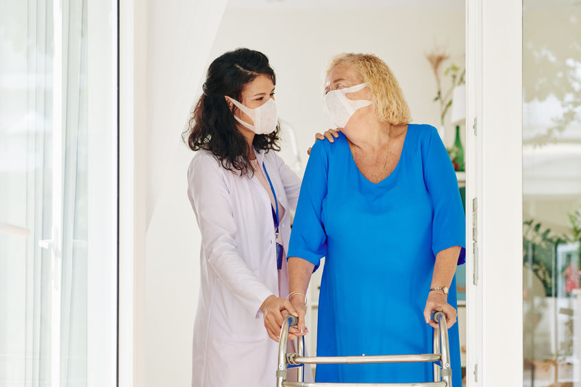 Caregiver in medical mask helping senior woman to move with help of walker